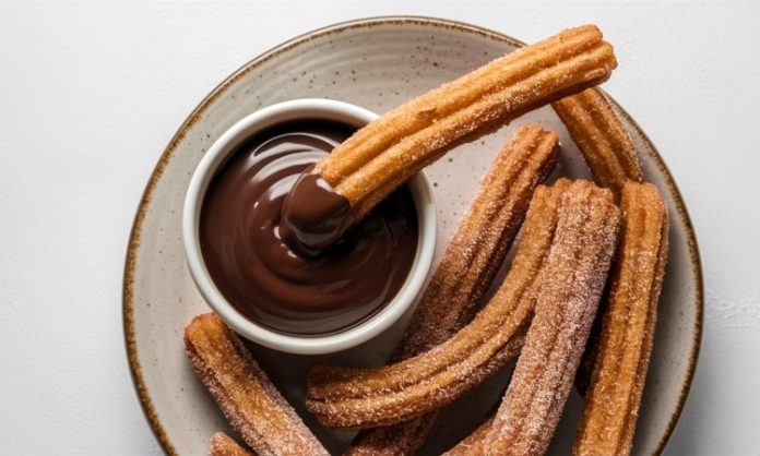 Churros Lezzetinin Yükselen Popülaritesi Tatlı Dünyasında Yeni Bir Dönem Churros, son yıllarda yalnızca İspanya’nın geleneksel sokak lezzeti olmaktan çıkıp dünya genelinde kafe menülerinin favorisi haline geldi. Özellikle Türkiye’de tatlı kültürünün giderek çeşitlenmesiyle birlikte churros popüler bir alternatif olarak öne çıkıyor. Çıtır yapısı, şeker ve tarçınla kaplanan dış yüzeyi ve sıcak çikolata sosuyla birleştiğinde ortaya çıkan benzersiz tat, birçok insanın yeni gözdesi haline gelmiş durumda. Konulu bir haber görseli.
